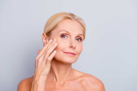 Mature woman applying facial cream, promoting skincare and beauty on a light background.