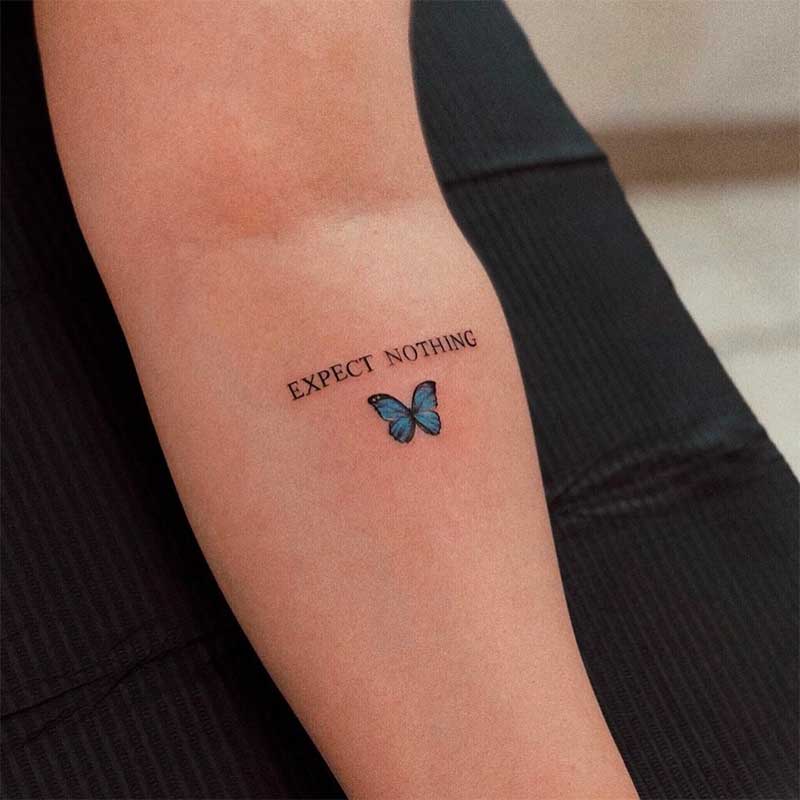 A "expect nothing" tattoo with a blue butterfly on the left forearm of a person with fair skin.