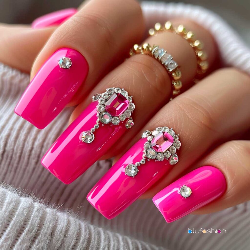 Luxurious hot pink coffin nails adorned with large pink and clear diamond rhinestones, creating an elegant and opulent manicure, complemented by gold stacked rings on the fingers.