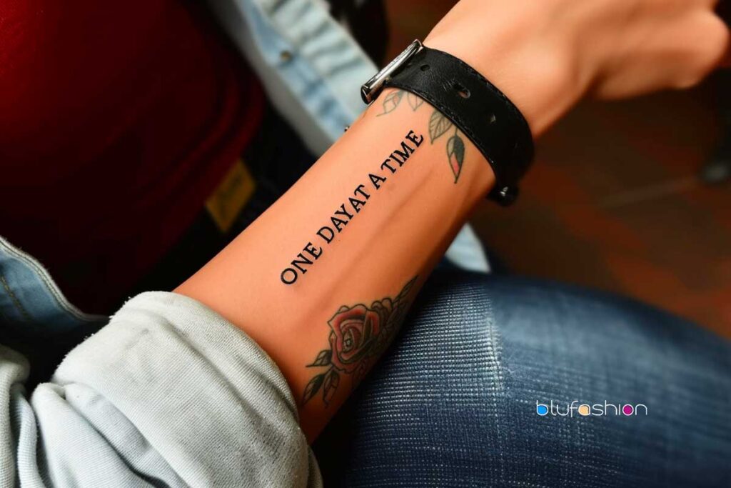 Inspirational 'One Day At A Time' tattoo in elegant script on inner forearm, paired with a watch and rose.