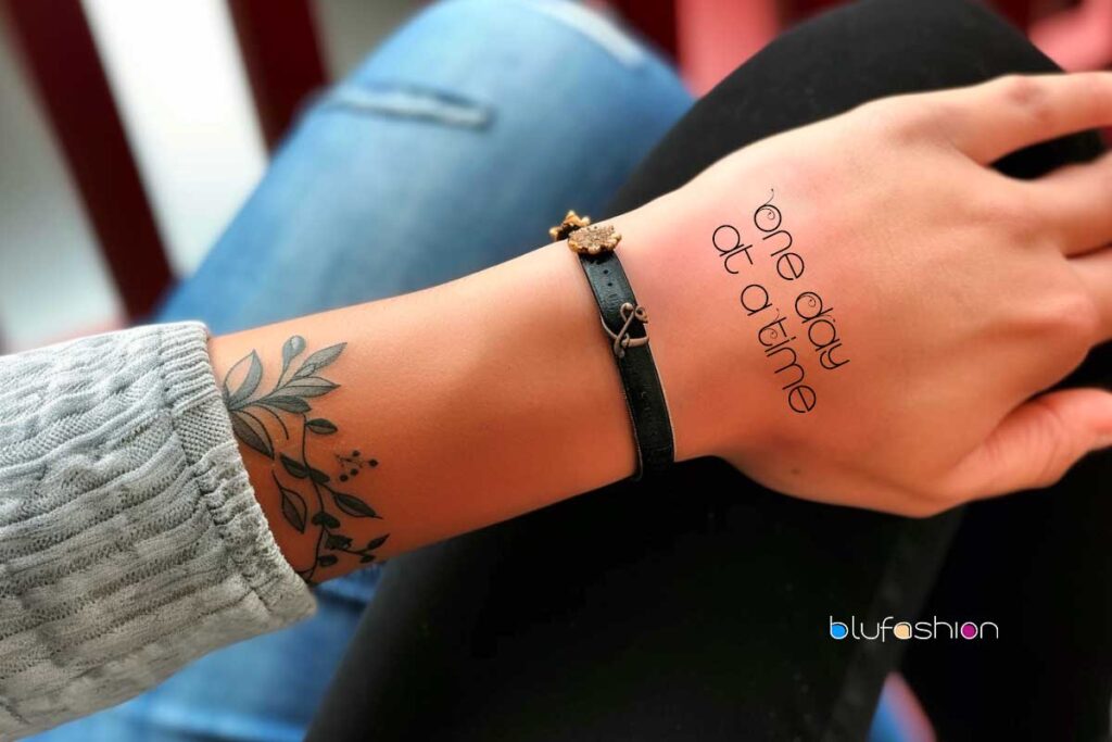 Simple 'One Day at a Time' cursive tattoo with floral design on a woman's forearm, showcasing mindfulness.