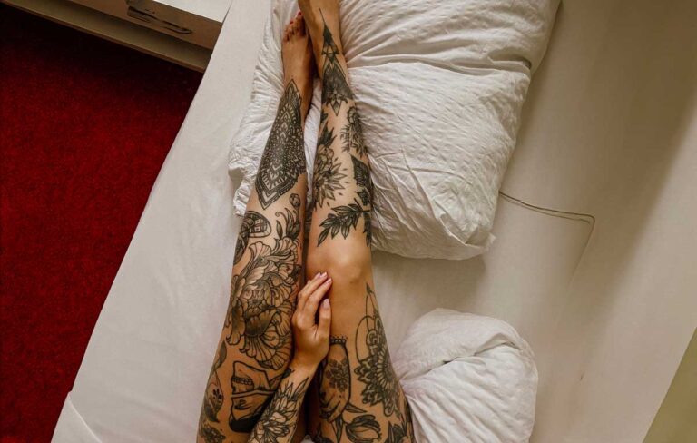 Intricately designed thigh tattoo on legs lying on a white bedspread with a red carpet background.