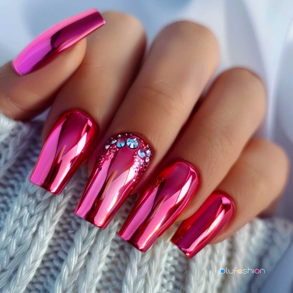 Shiny metallic hot pink coffin nails with an accent nail featuring a decorative arc of blue and clear rhinestones at the base, set against a cozy grey knit background.